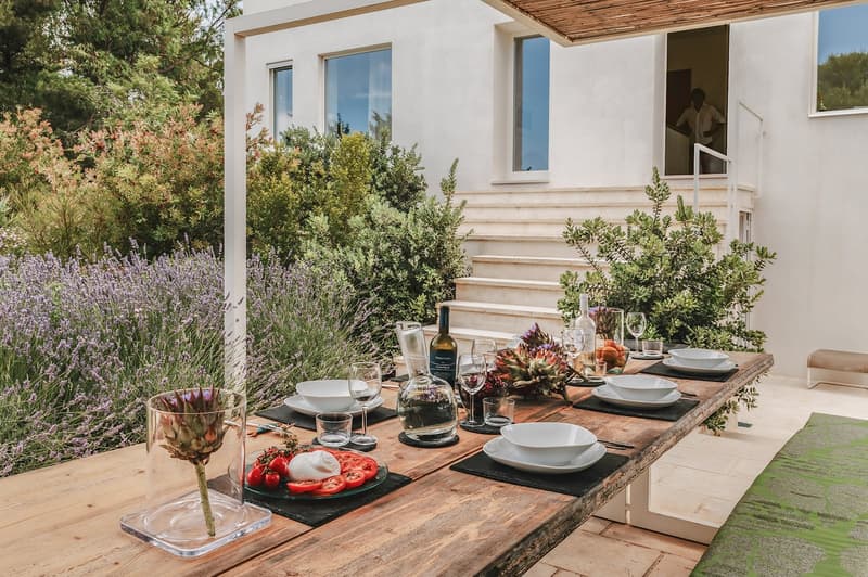 A wooden table set for a meal with a view of a white house with stairs and a garden in the background.