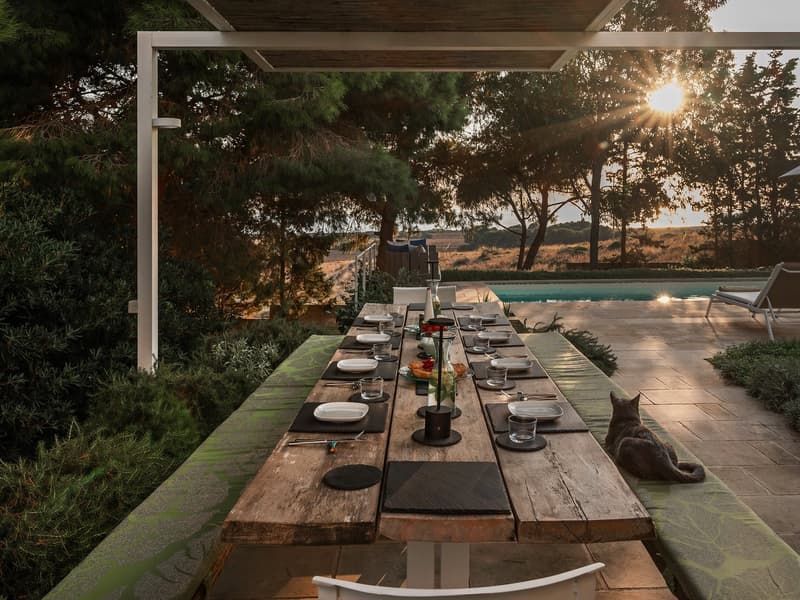 Long wooden table, set for dining, outdoor dining area, swimming pool in the background, trees, sun setting