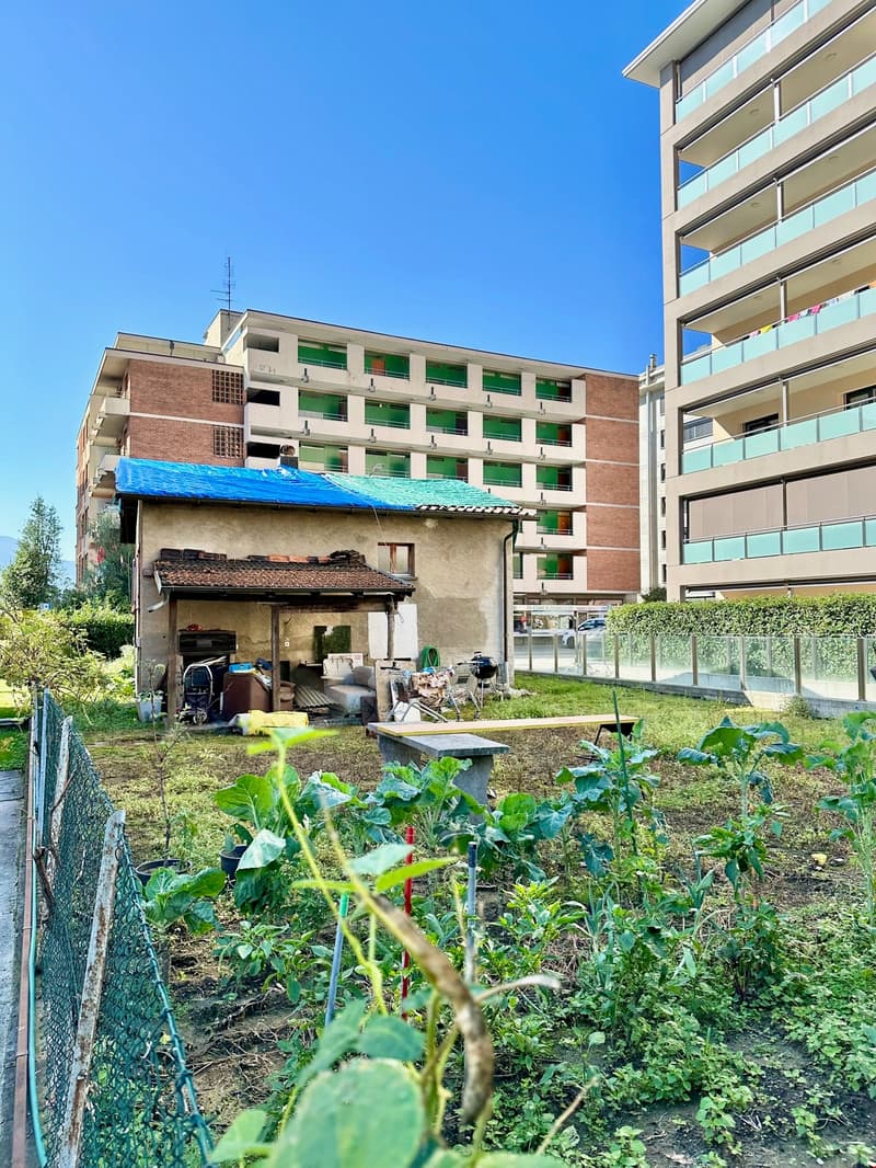 A small house with a garden, two larger apartment buildings in the background, bicycle, chairs, table and some storage boxes.