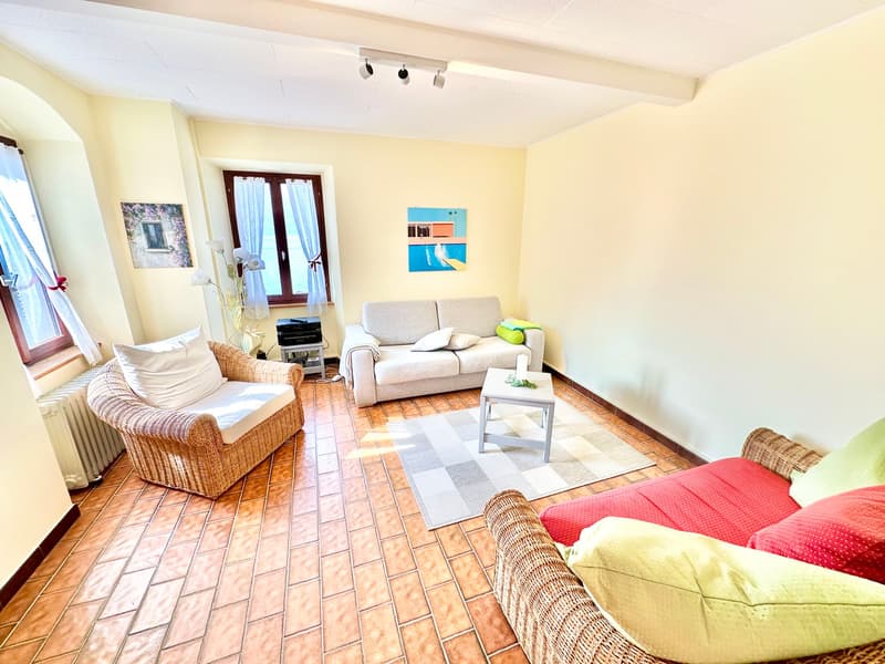 Living room with beige walls, brown tile floor, white ceiling, two couches, coffee table, rug, radiator, windows with white curtains, picture frames, and decorative items.
