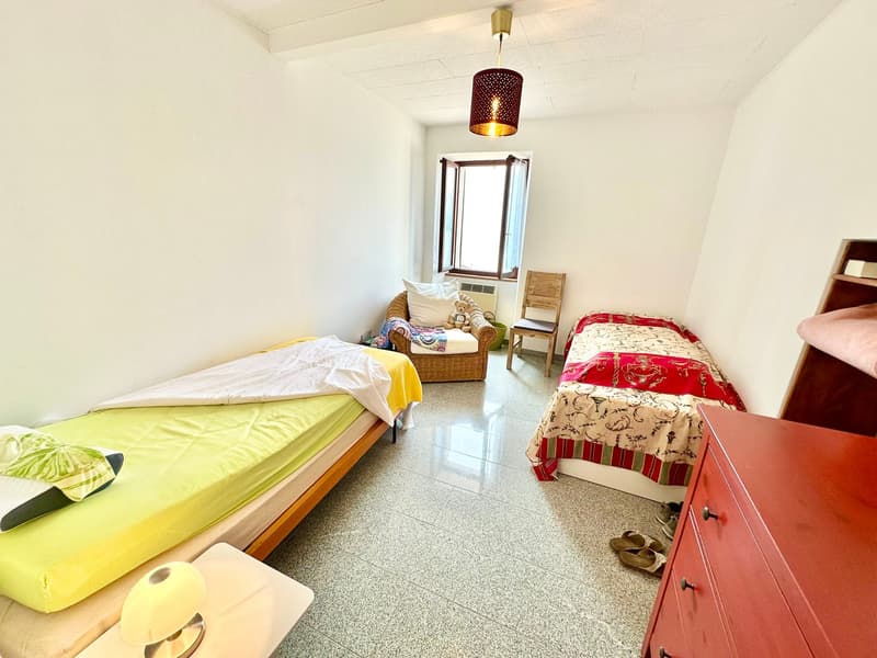 Two single beds, a chair, a wicker chair with a pillow, a window, a lamp, a red cabinet, a shelf, and a lamp on a table.