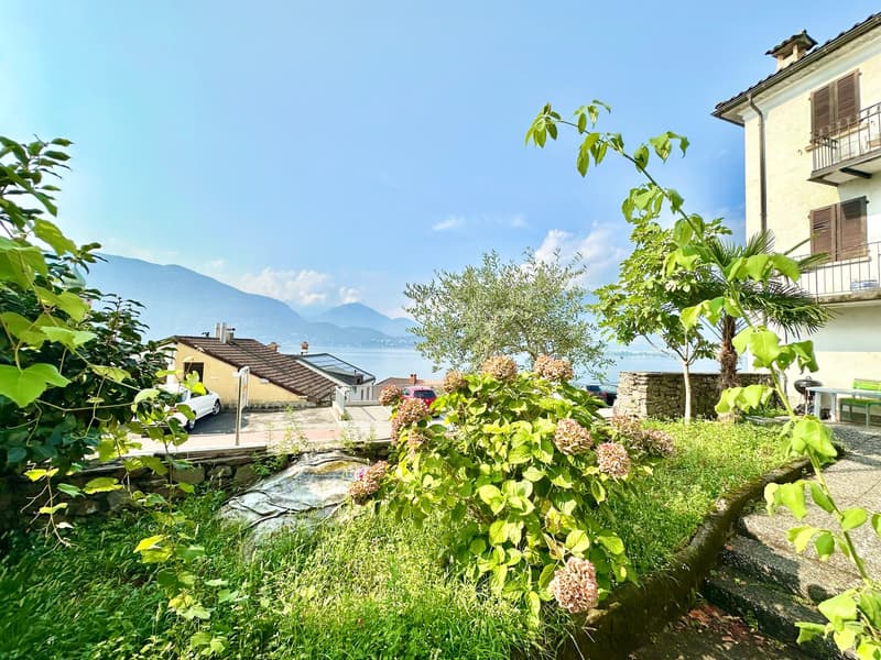 Garden with plants and trees, view of a lake and mountains in the background, a house with a balcony
