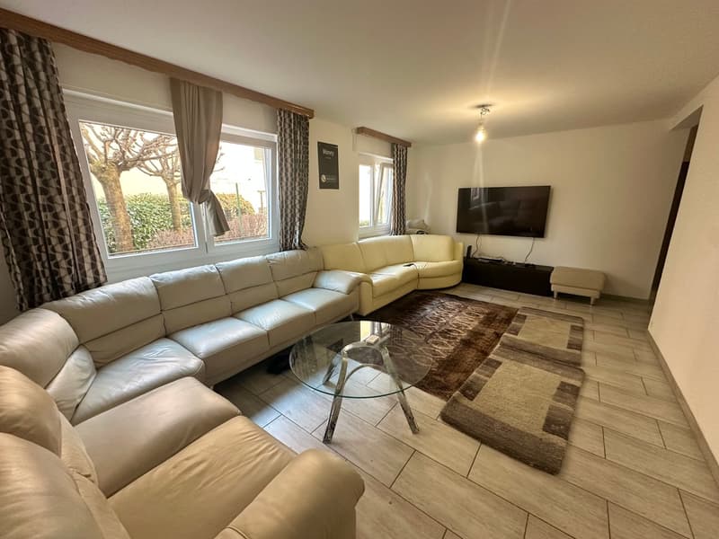 Spacious living room with large sectional sofa, glass coffee table, TV, and patterned area rug. The room has large windows overlooking what appears to be a balcony or terrace.