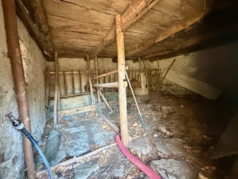 The image shows an interior of an abandoned building or structure. The ceiling is made of wooden beams and the walls are made of stone or concrete. There are various debris and discarded items scattered on the floor, including a metal frame and a pink hos