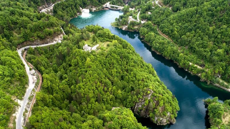The image shows a lush, green landscape with a winding river or lake running through it. The area is surrounded by dense forests and rocky cliffs, creating a picturesque natural setting. There appears to be a road or path running along the edge of the wat