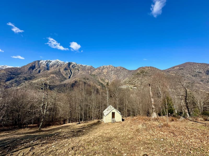 The image shows a picturesque mountain landscape with snow-capped peaks in the background. In the foreground, there is a small, rustic stone building surrounded by a bare, grassy field and a dirt path. The scene is framed by a forest of bare trees on eith