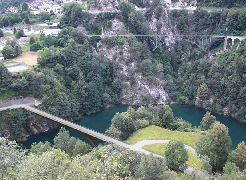 The image shows a scenic mountain landscape with a winding river or lake below. There is a large bridge spanning the valley, surrounded by dense forest and rocky cliffs. The landscape appears to be in a mountainous, forested region.