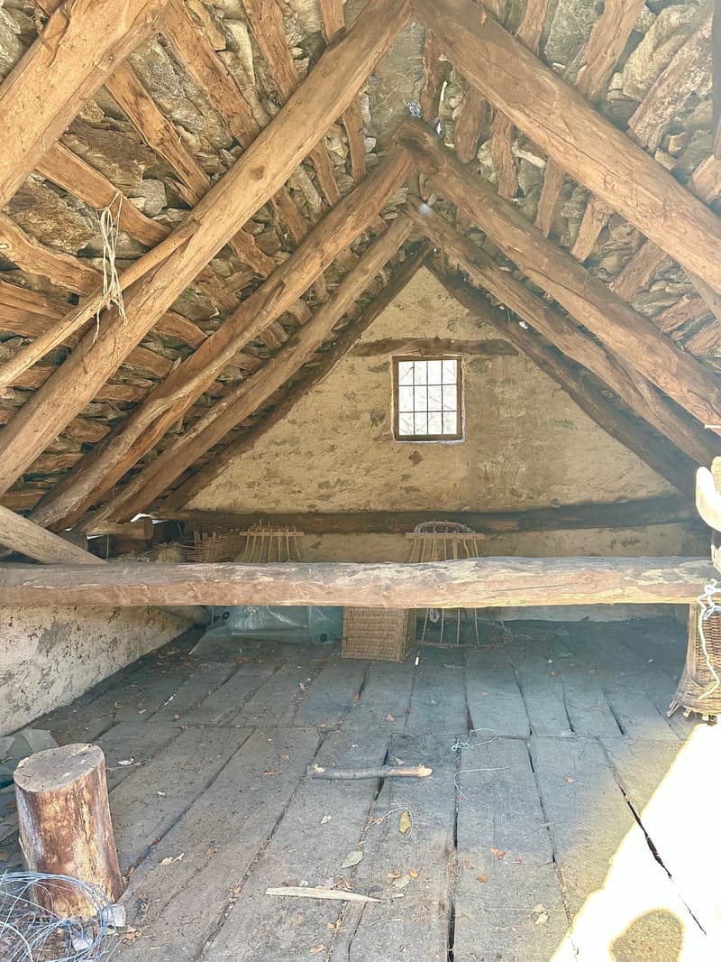 The image shows the interior of an attic space. The ceiling is made of exposed wooden beams and rafters, creating a rustic and traditional appearance. The floor is covered with wooden planks, and there is a small window that allows natural light to enter
