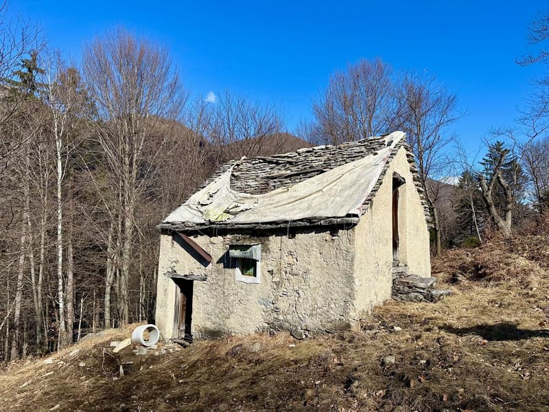 An old, dilapidated stone cabin with a slanted, slate roof in a forested, mountainous area. The cabin appears to be abandoned, with a small garage or outbuilding visible next to it.