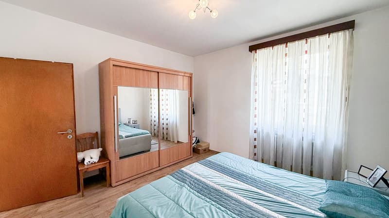 The image shows a spacious bedroom with a large wooden wardrobe, a bed with blue bedding, and a window with white curtains. The room has a warm and cozy feel with the wooden furniture and neutral color scheme.