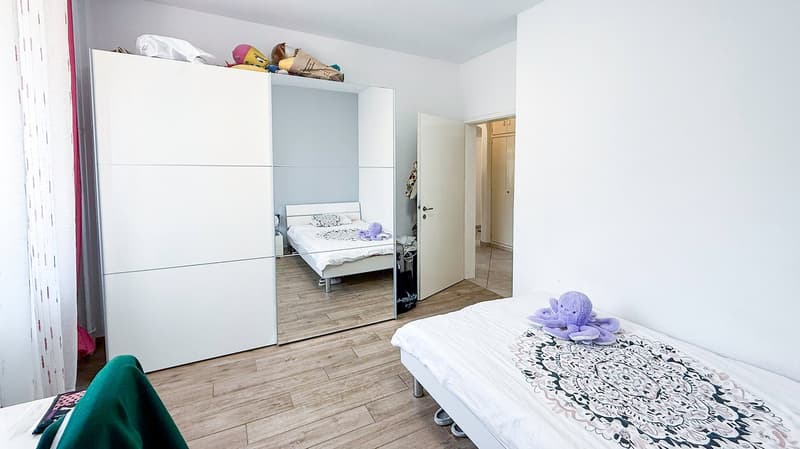 Spacious bedroom with a white wardrobe, a bed with a patterned comforter, and a plush purple stuffed animal. The room has hardwood flooring and white walls.