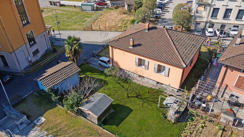 Two-story residential building with a red tiled roof, surrounded by a grassy area and trees. There is a small garden or yard in the foreground, and a parking area visible in the background.