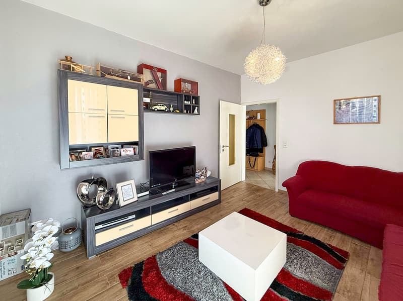 Spacious living room with hardwood flooring, gray walls, a large TV stand with shelves, a red sofa, a white coffee table, and a decorative chandelier. The room also features a balcony or terrace and a fireplace.