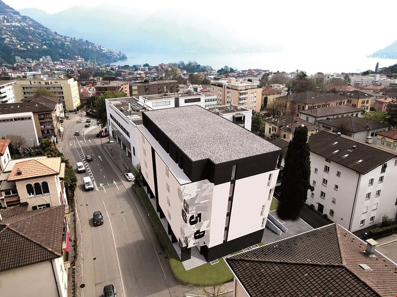 Large modern multi-story building with a flat gray roof, surrounded by other residential and commercial buildings in a hilly urban area with a view of the mountains in the background.