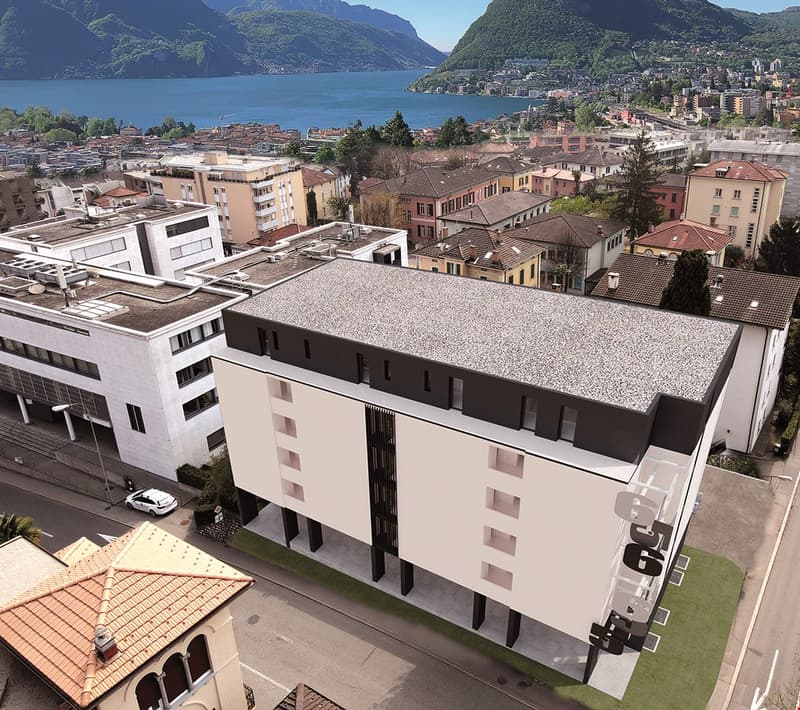 Multi-story residential buildings with gravel roofs, surrounded by a lake and mountains in the background. The buildings appear to be located in a city or town setting.