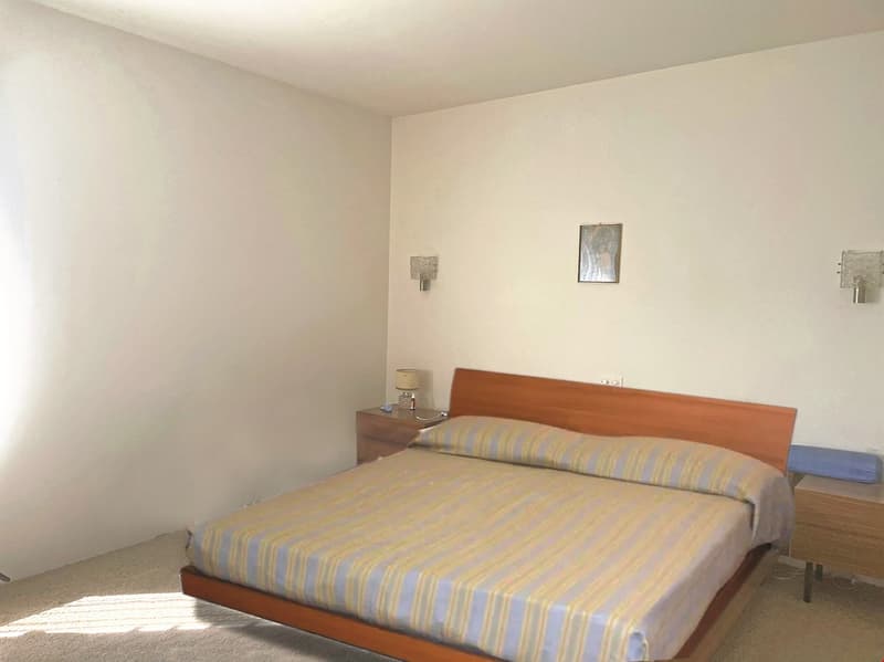 The image shows a bedroom with a wooden bed frame, a striped bedsheet, and a small nightstand. The walls are painted in a light, neutral color, and there is a framed artwork hanging on the wall.