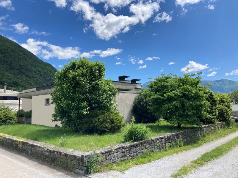 The image shows a scenic outdoor landscape with a grassy area, lush green trees and shrubs, and a stone wall surrounding the property. The sky is blue with fluffy white clouds, creating a picturesque natural setting.