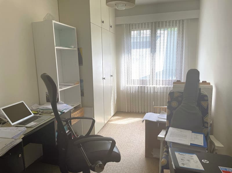 Bedroom with a desk, laptop, and storage cabinets. Large window with curtains overlooking a balcony or terrace.