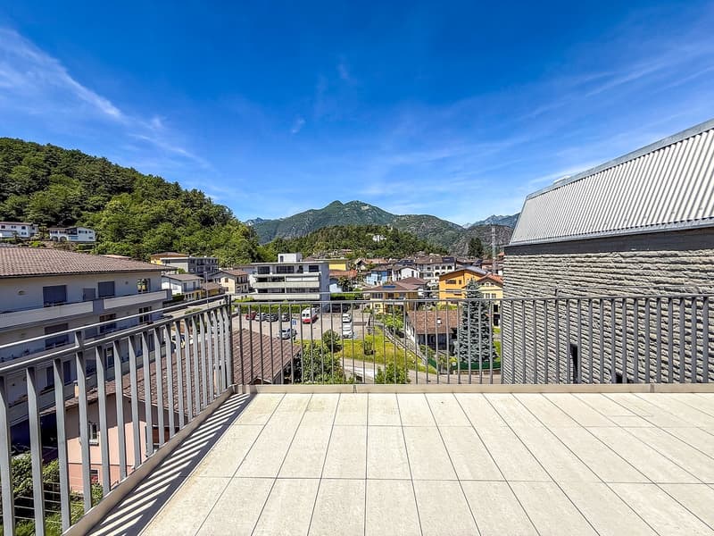 The image shows a balcony or terrace overlooking a scenic mountain town. The balcony has a tiled floor and metal railings. In the distance, there are colorful buildings and lush green trees against a backdrop of mountains and a clear blue sky.