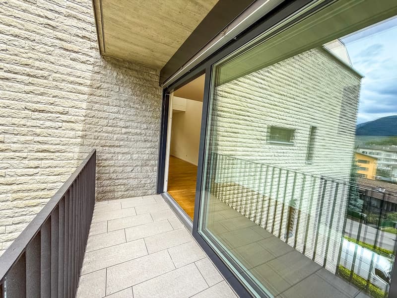 The image shows a balcony or terrace area with a stone or tile floor, large glass windows, and a stone or brick exterior wall. The balcony appears to have a railing and offers a view of the surrounding landscape.