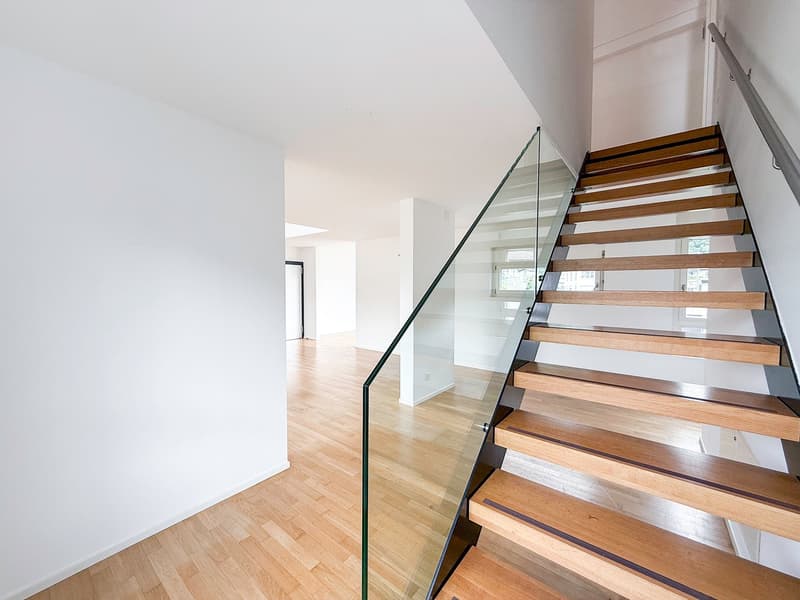 Spacious open-plan living area with hardwood floors, glass staircase with wooden steps, and large windows providing natural light.