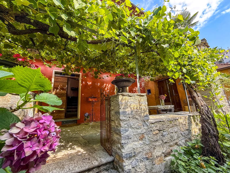 Rustic stone house with a covered patio, surrounded by lush greenery and colorful flowers. The house has a stone wall and a decorative fountain or birdbath. There are large windows and a wooden door leading into the house.
