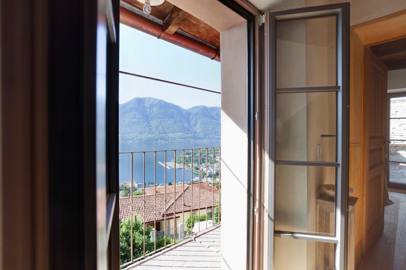 Spacious room with large windows offering a stunning view of the mountainous landscape and the town below. The room features wooden beams and a balcony or terrace overlooking the scenic vista.