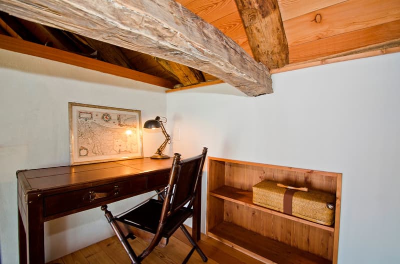 The image shows a cozy home office space with exposed wooden beams on the ceiling, a wooden desk with a lamp, and a wooden bookshelf. The room has a rustic, cabin-like feel with the wooden elements and the framed artwork on the wall.