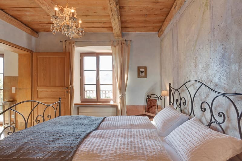 Rustic bedroom with wooden ceiling beams, a crystal chandelier, a balcony with wrought iron railings, and a fireplace. The room features a wrought iron bed frame and bedding in neutral tones.