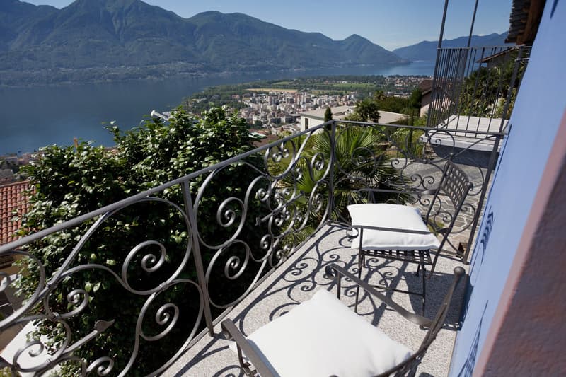 Ornate wrought-iron balcony railing, lush greenery, and a panoramic view of a lakeside town surrounded by mountains