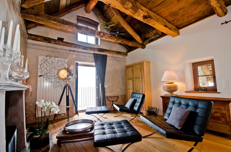 Rustic living room with exposed wooden beams on the ceiling, leather armchairs and ottoman, a fireplace, and decorative lighting fixtures. The room has a cozy and warm atmosphere with a mix of modern and traditional elements.
