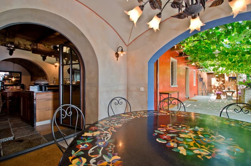 Cozy indoor dining area with arched doorway leading to an outdoor patio or balcony area. The dining table has a colorful floral mosaic top. The room features warm lighting, wrought iron chairs, and a decorative overhead light fixture. The exterior visible
