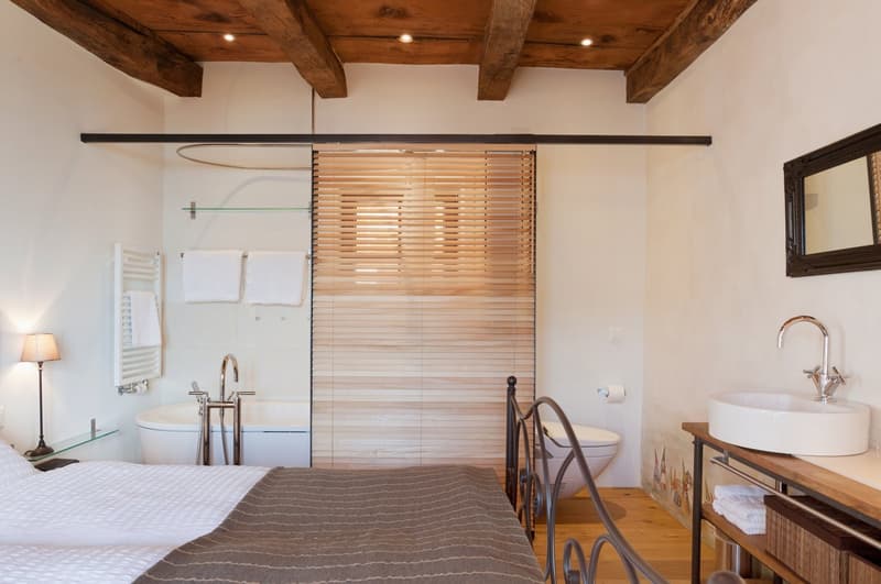 The image shows a spacious bedroom with an en-suite bathroom. The bedroom features a wooden ceiling with exposed beams, a large bed with a gray striped comforter, and a sliding wooden partition that separates the bathroom area. The bathroom includes a toi