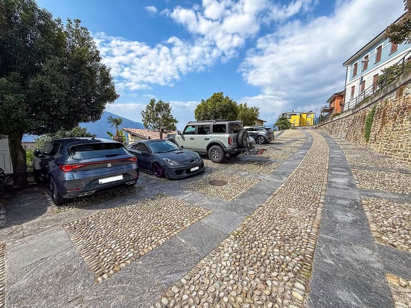 The image shows a paved outdoor area with a cobblestone path, surrounded by trees and buildings. There are several vehicles parked in the area, including a black SUV and a sports car. The sky is blue with some clouds, and the overall scene appears to be i