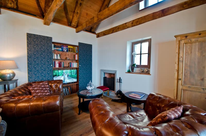 Spacious living room with high wooden beamed ceiling, leather sofas, wooden shelves with books, and a fireplace. The room has a cozy and rustic feel with the exposed wooden beams and warm lighting.