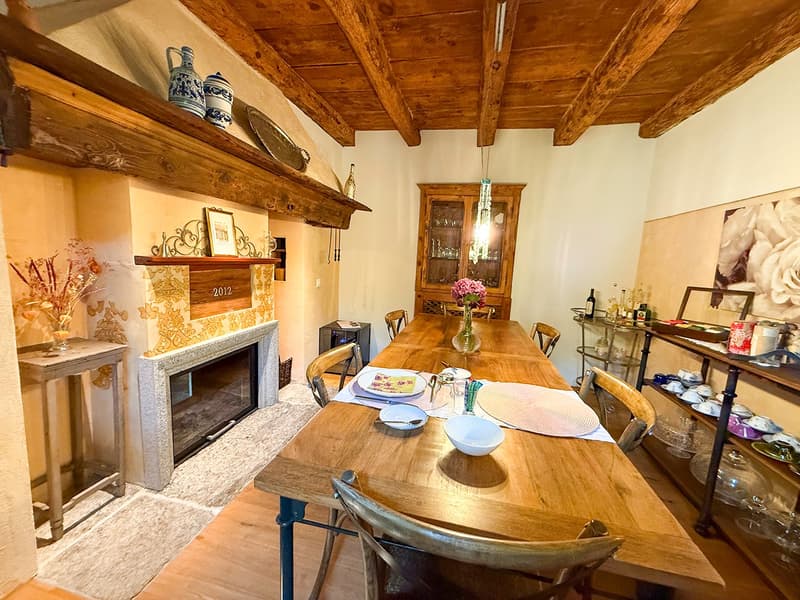 Rustic dining room with wooden beamed ceiling, wooden dining table and chairs, fireplace, shelves with decorative items, and a cabinet with glassware.