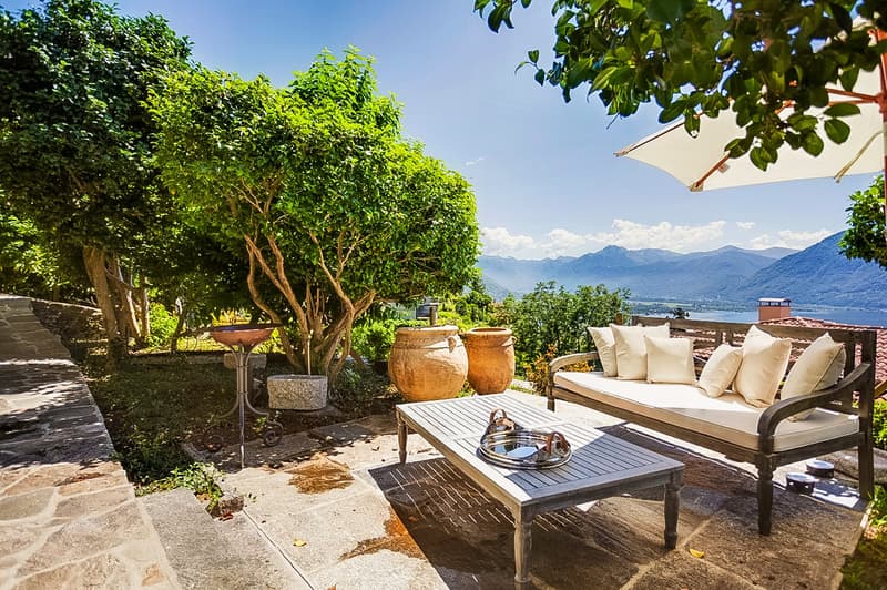 Lush green trees and foliage, stone patio with wooden furniture, large outdoor umbrella, mountain view in the background