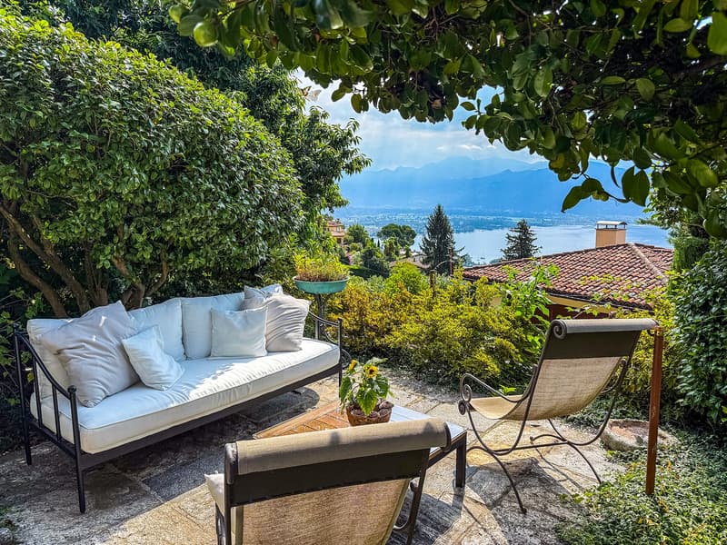 Cozy outdoor patio with a comfortable sofa, chairs, and a scenic view of the lake and mountains in the background, surrounded by lush greenery and trees