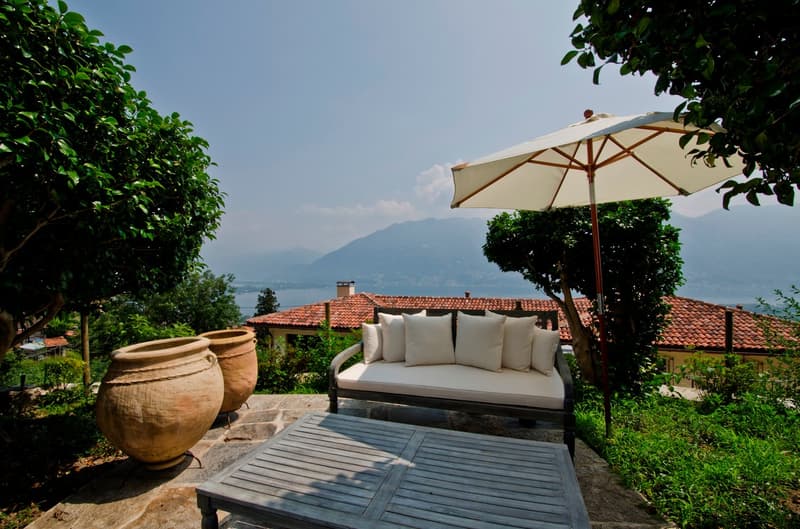 The image shows an outdoor patio area with a comfortable sofa, a large umbrella, and two large terracotta pots. The patio overlooks a scenic landscape with mountains in the background, indicating a beautiful view. The patio is surrounded by lush greenery,