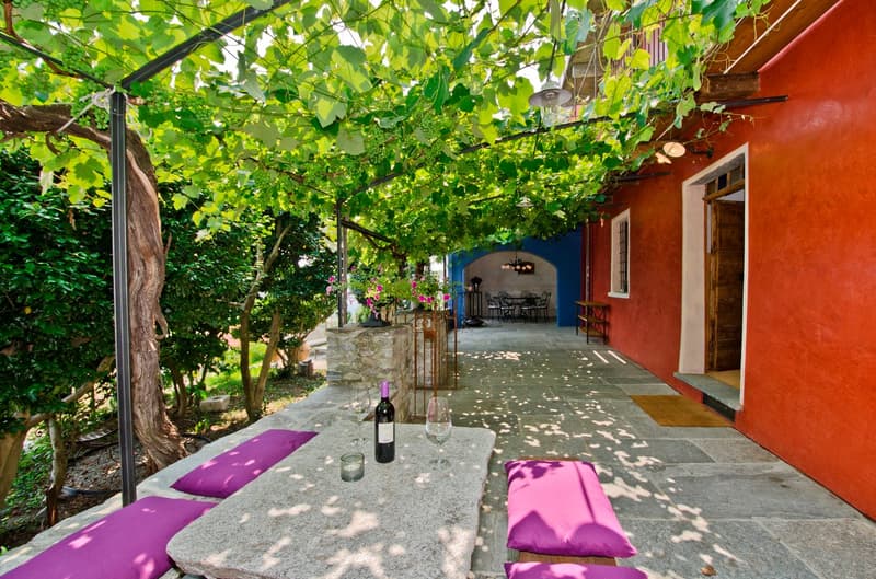 Covered outdoor patio with stone floor, red brick walls, and a wooden trellis covered in lush green vines. There is a table with wine glasses and a bottle of wine, as well as purple cushions on the seating area.