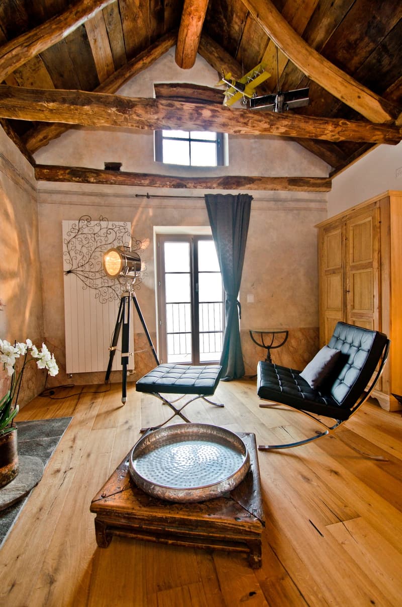 Rustic living room with exposed wooden beams, large windows, leather armchair, decorative tripod lamp, and a wooden coffee table with a decorative bowl on it.