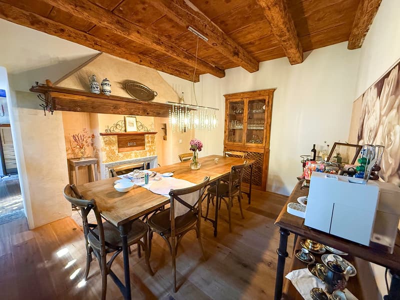 Rustic dining room with wooden beamed ceiling, wooden dining table and chairs, fireplace, shelves with decorative items, and a glass-door cabinet