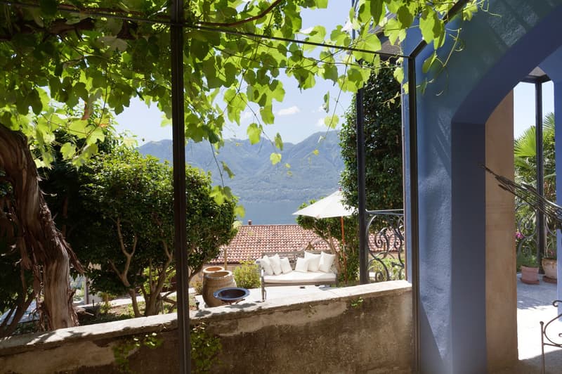 Covered outdoor patio with a couch, surrounded by lush greenery and trees. The patio overlooks a scenic lake and mountains in the distance.