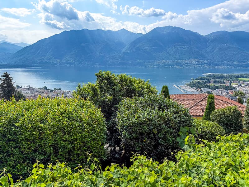 The image shows a scenic view of a mountainous landscape with a large lake in the foreground. The mountains are surrounded by lush, green vegetation, and the lake is dotted with a marina or harbor. The sky is partly cloudy, creating a picturesque and sere