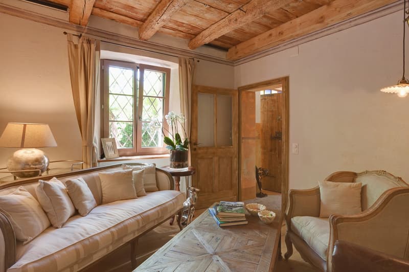 Cozy living room with wooden beamed ceiling, large windows, plush beige sofa and armchair, wooden coffee table, and a fireplace