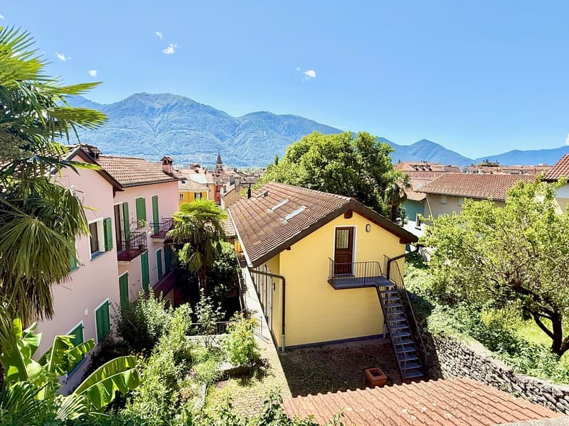 Terraced house, yellow exterior, brown roof, balcony, stairs, green windows, trees, mountains in background