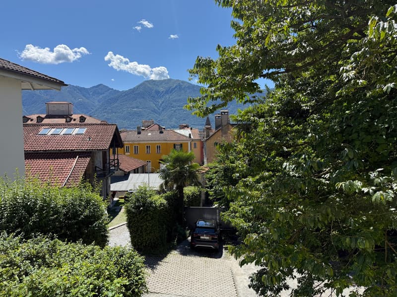 multi-story buildings, red roofs, mountains in the distance, driveway, black car parked, trees and bushes, paved pathway