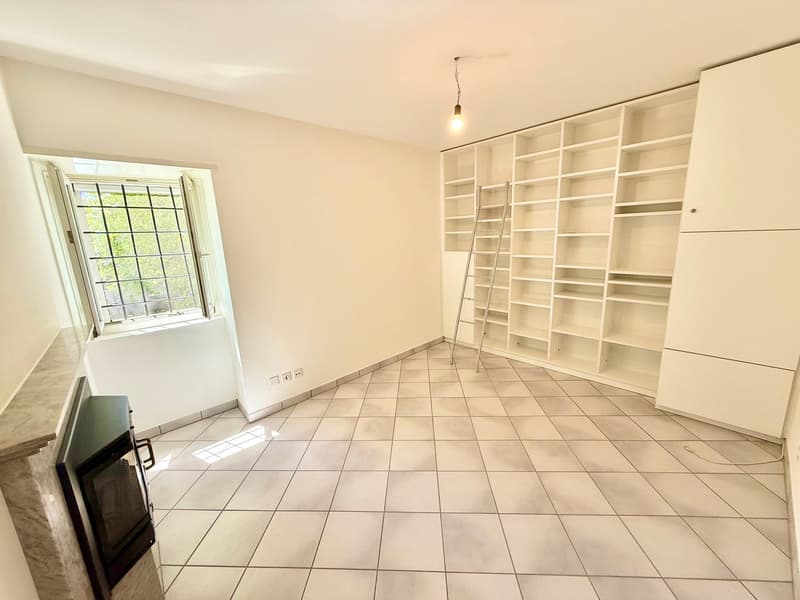 empty room with white tiled floor, white walls, small fireplace, full-length built-in shelves, window with outdoor view, pendant light, ladder, and power outlets
