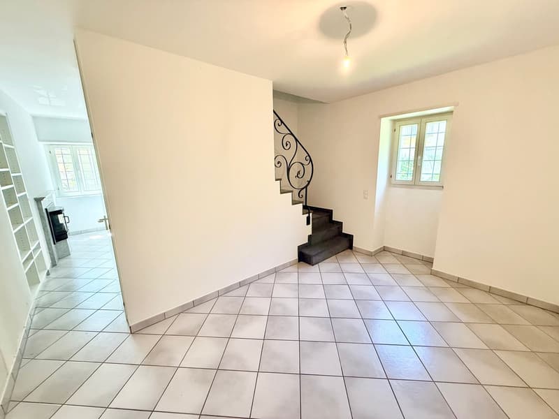 Empty room with white tiles, white walls, wooden floor under stairs, stairs with metal railing, small window with white frames, ceiling lamp