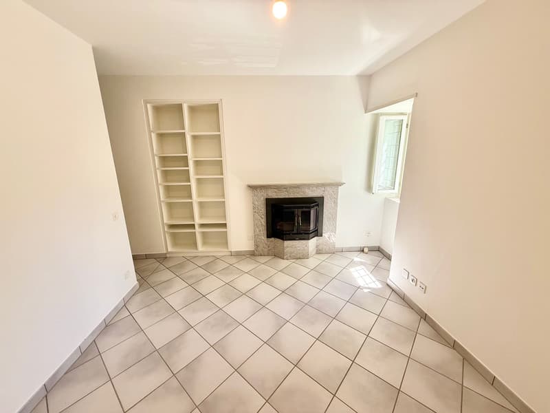 Empty living room with white walls, tiled floor, built-in fireplace, shelf, and a window on the right side.